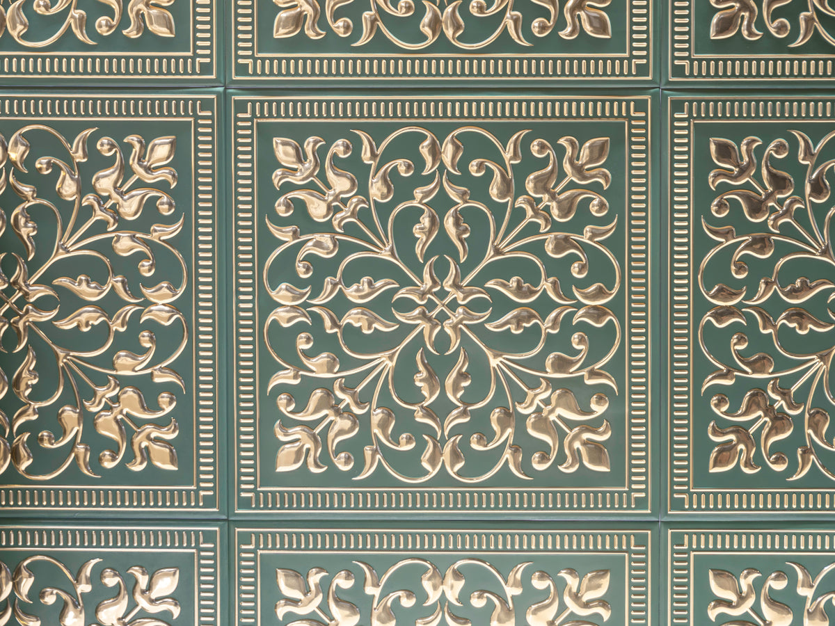 Close-up of embossed metal tiles with bright gold scrollwork and beaded borders pressed into a deep green ground, laid edge to edge in a grid