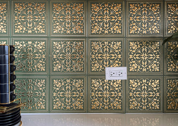 Dark green peel and stick metal tiles with gold scrollwork sitting flush on a kitchen backsplash around a white outlet, with a stack of black mugs on the counter