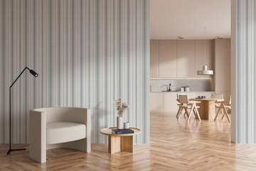 Blue-grey striped wallpaper on two walls framing an opening into a pale kitchen and dining area, with a cream armchair, black floor lamp and round wood side table on a herringbone floor