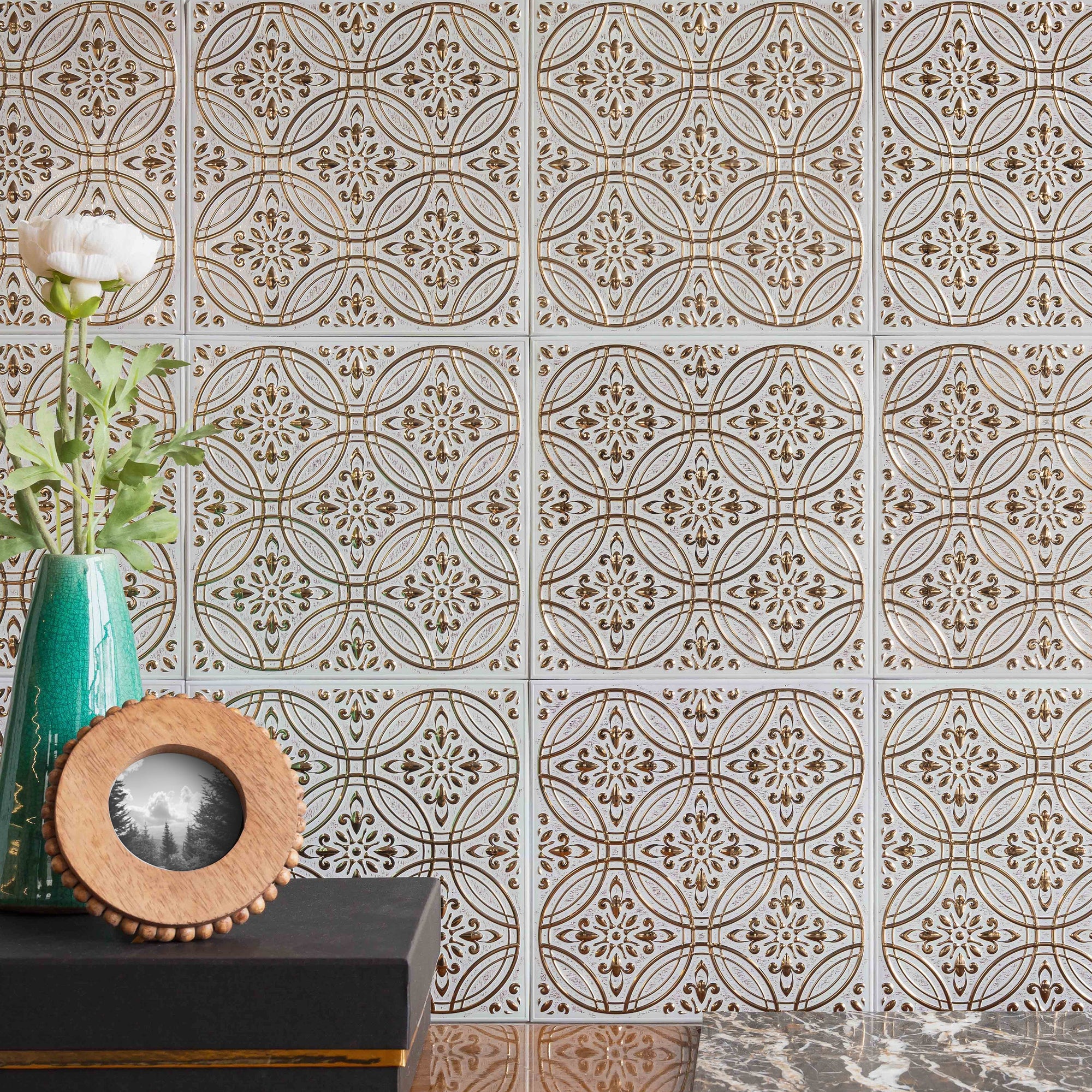 A continuous field of white and gold embossed metal tiles with the seams visible, behind a dark console holding a teal vase of white ranunculus and a small round wooden picture frame.