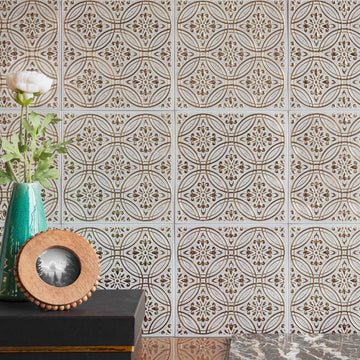 A continuous field of white and gold embossed metal tiles with the seams visible, behind a dark console holding a teal vase of white ranunculus and a small round wooden picture frame.