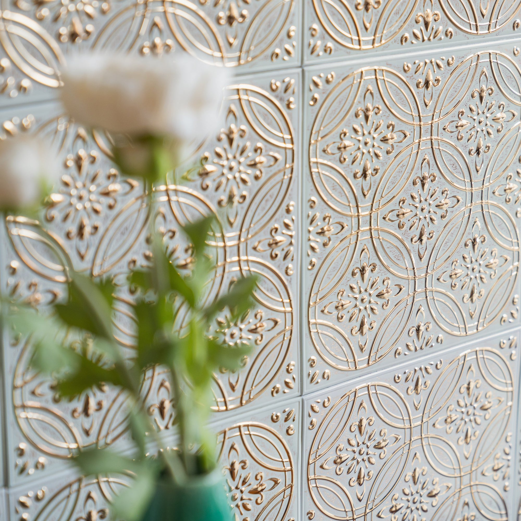 Close-up of aged white and gold embossed metal tiles on a wall in low angled daylight, with white ranunculus in a teal vase blurred in the foreground