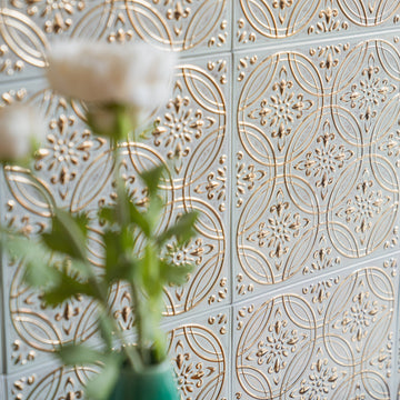 Close-up of aged white and gold embossed metal tiles on a wall in low angled daylight, with white ranunculus in a teal vase blurred in the foreground