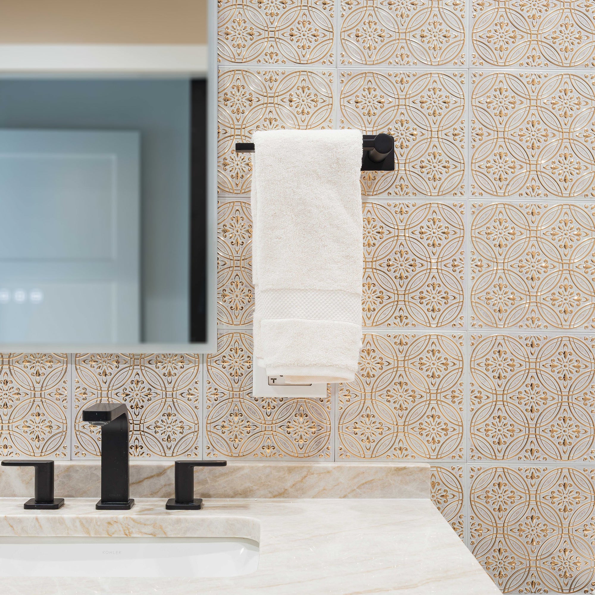 Ornate gold and white embossed metal tiles covering a bathroom vanity wall, with a black faucet, marble countertop and a framed mirror.