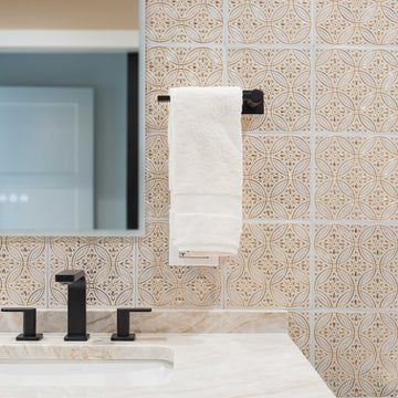 Ornate gold and white embossed metal tiles covering a bathroom vanity wall, with a black faucet, marble countertop and a framed mirror.