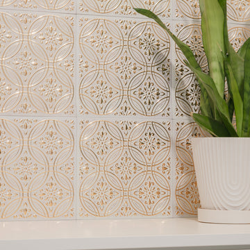 A wall of antique white-and-gold peel-and-stick metal tiles in a pressed-tin medallion pattern, styled as an accent behind a shelf with a potted plant — the tin ceiling look in real aluminum.