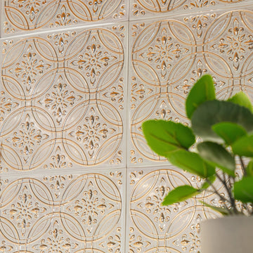 Whitewashed gold peel and stick metal tiles seen at an angle, sunlight catching the embossed relief, with glossy green leaves at the right edge.