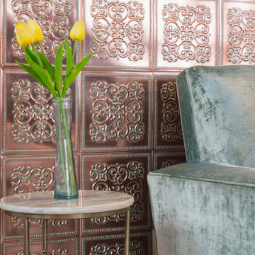 Copper-finish pressed metal tiles with scrollwork and beaded borders covering a wall behind a velvet armchair and a marble side table with yellow tulips