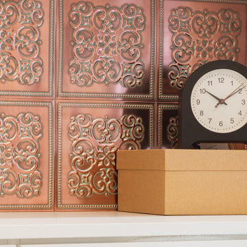 Copper-toned metal tiles with ornate scrollwork relief covering a wall behind a white shelf, with a black desk clock and a kraft storage box.