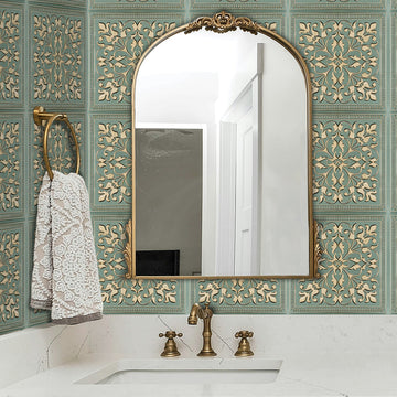 Vanity backsplash in sage green metal tile with raised cream and gold scrollwork, behind an undermount sink, antique brass faucet and an arched brass-framed mirror