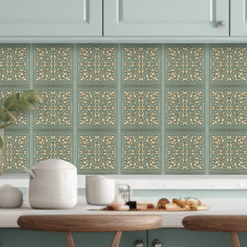 Rustic Farmhouse peel and stick metal tiles, gold ornamental relief on a soft green ground, installed as a kitchen backsplash between the countertop and sage-green upper cabinets, with white ceramic crocks and a wooden board of snacks on the counter