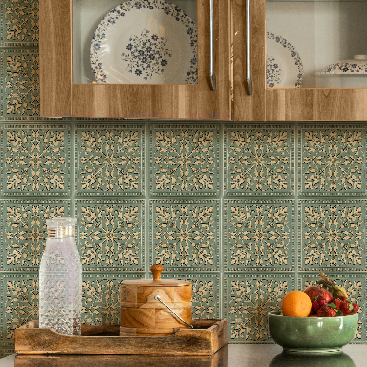 Sage-green and cream patterned metal backsplash tiles on a kitchen wall beneath warm wood glass-front cabinets, with a dark countertop, fruit bowl and wooden tray below.