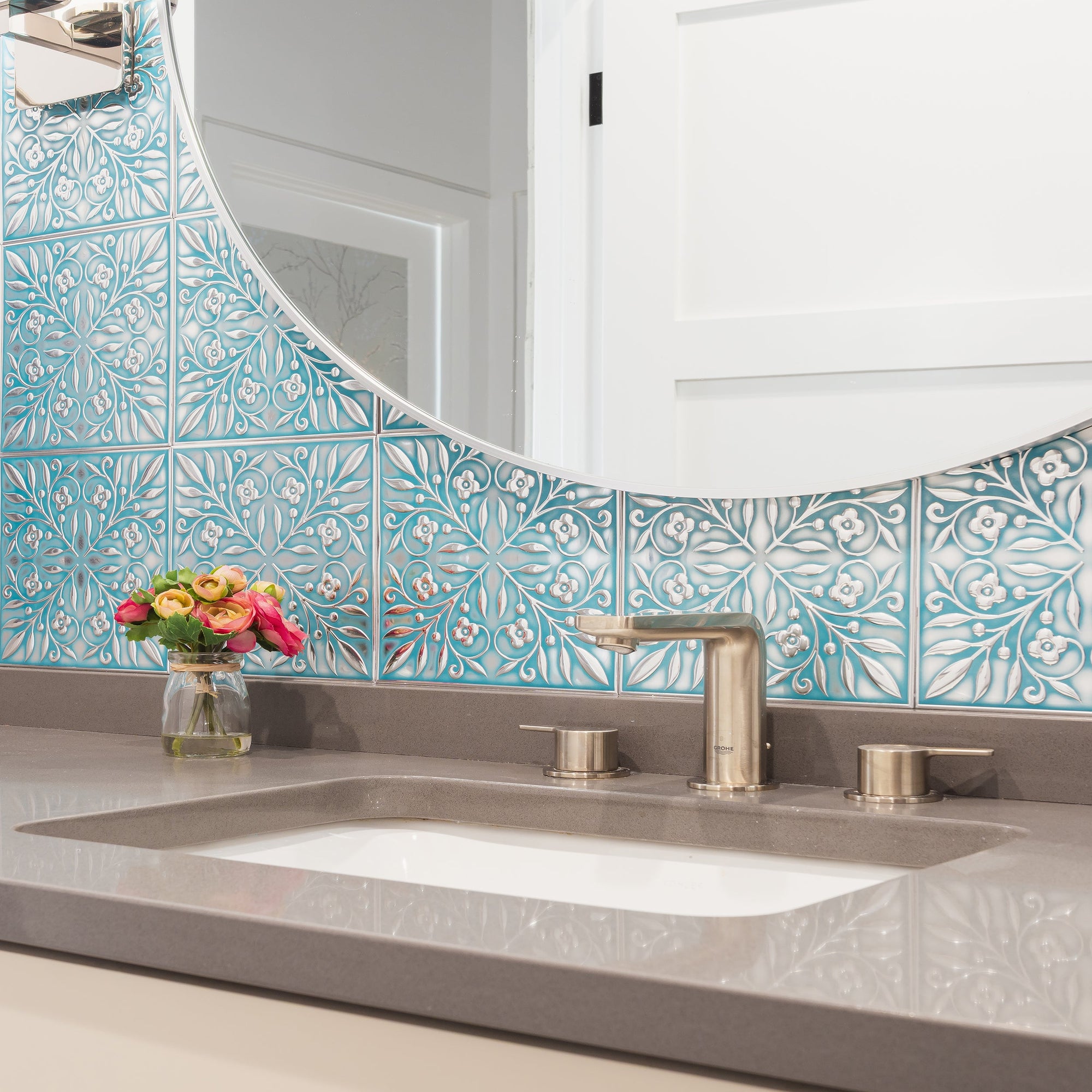 Teal and silver leaf-patterned metal tiles laid as a small-format grid on a bathroom vanity backsplash, with a round mirror, brushed faucet and a jar of flowers.