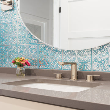 Teal and silver leaf-patterned metal tiles laid as a small-format grid on a bathroom vanity backsplash, with a round mirror, brushed faucet and a jar of flowers.