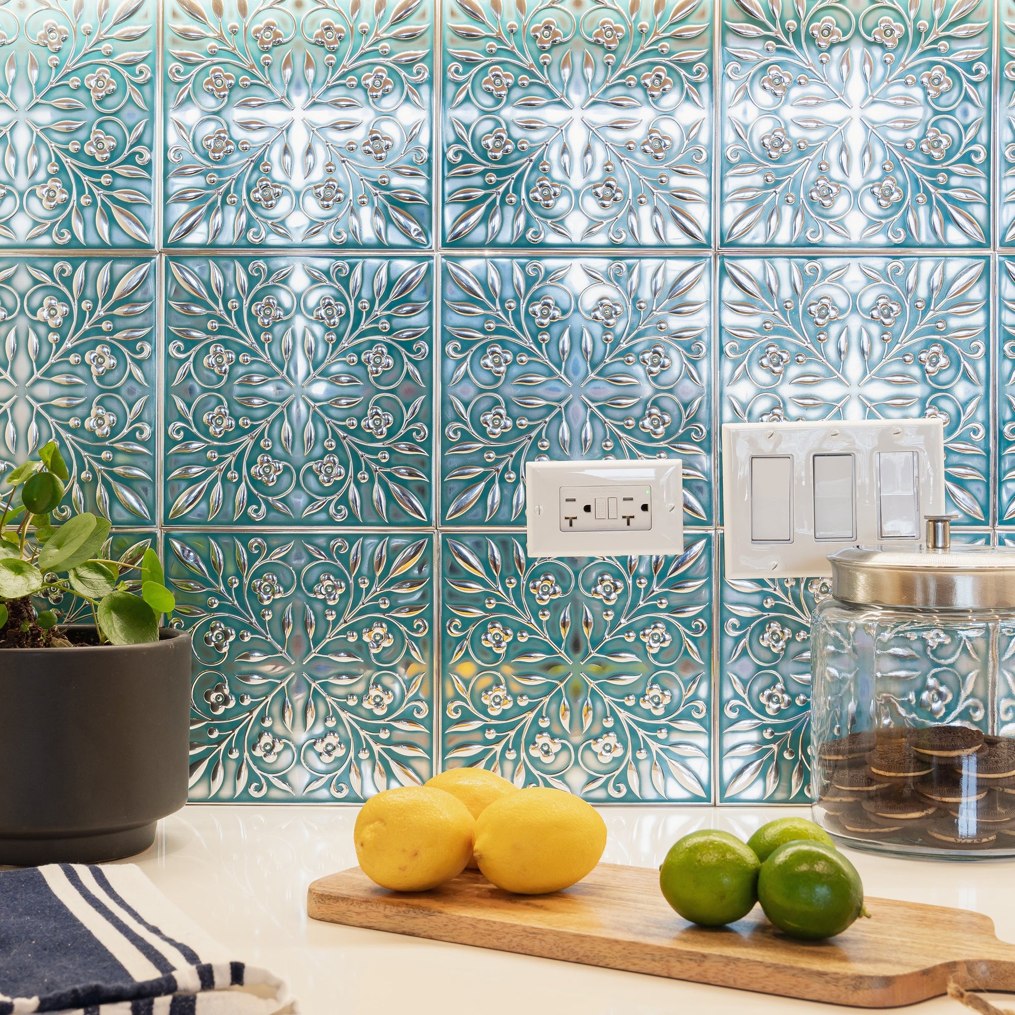 Teal and Silver Leaves peel and stick metal tiles installed as a kitchen backsplash around an outlet and light switches, with lemons and limes on the counter