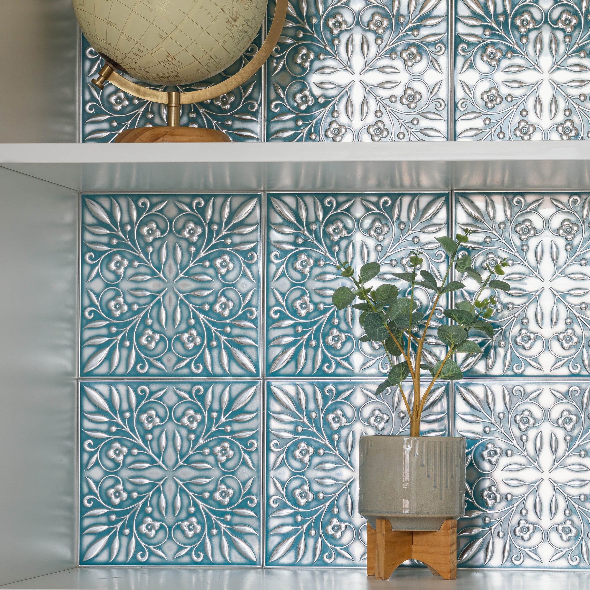 Teal and silver embossed metal tiles behind a shelf holding a globe and eucalyptus, showing the two-tone finish people aim for when painting tin ceiling tiles.