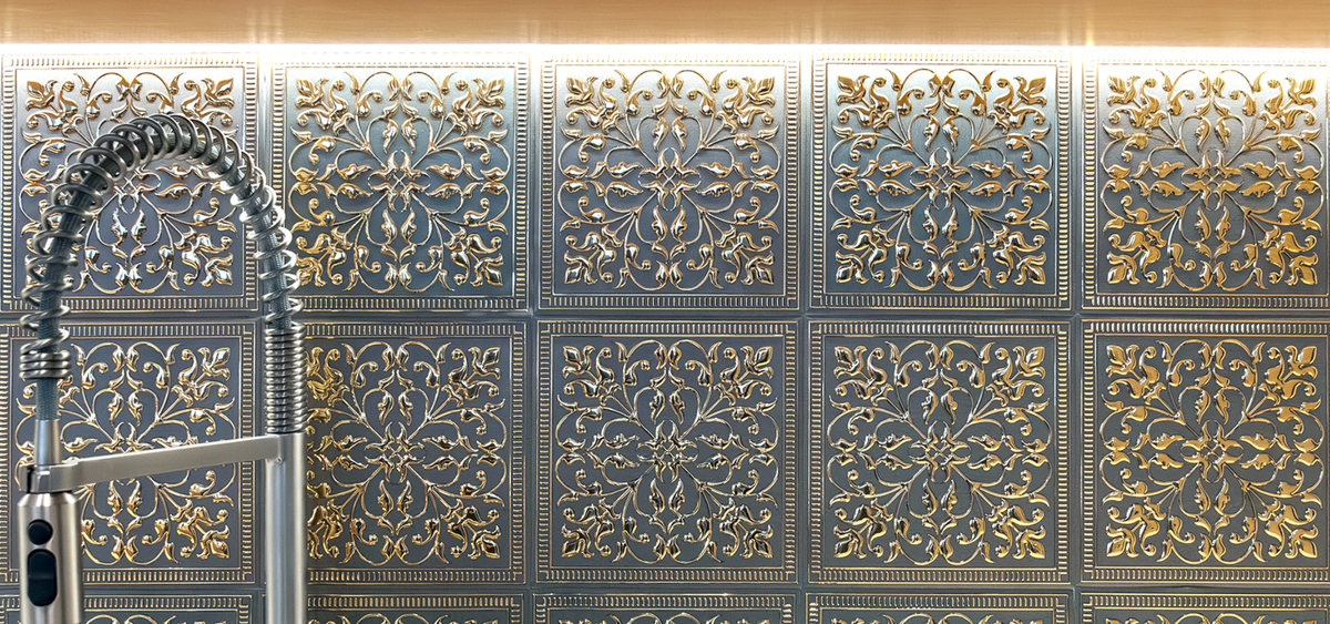 Rows of square embossed metal tiles with gold scrollwork on a dark silver ground under warm under-cabinet lighting, behind a stainless pull-down kitchen faucet