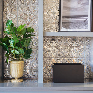 Silver Fleur embossed metal tiles lining the back of a white shelving unit, showing the pressed-tin medallion pattern with a plant and framed print.