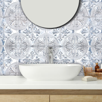 Silver metal tiles with an ornate floral pattern behind a white vessel sink on a light oak vanity, with a round mirror and amber toiletry bottles