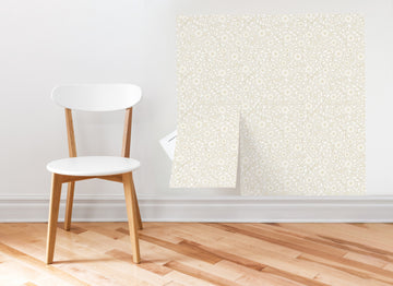 A Plot Thicken peel and stick wallpaper sheet mid-application on a wall beside a wooden chair, showing the small-format sheet being smoothed into place