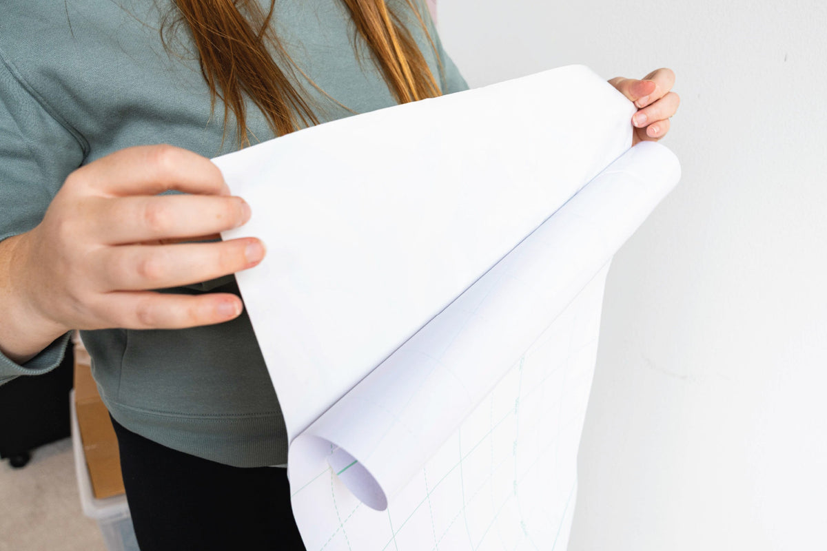 Hands holding a roll of peel and stick wallpaper with its white, grid-printed backing liner curling away from the paper