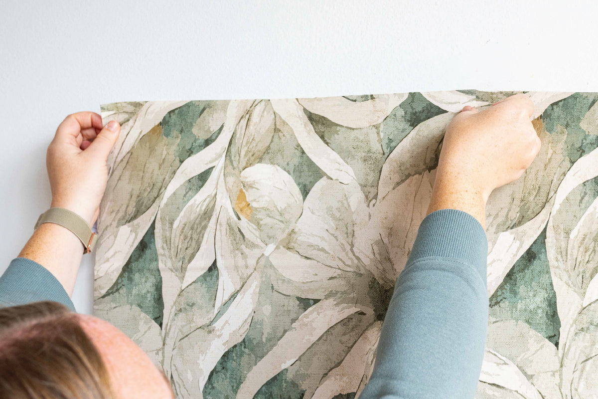 Two hands holding the top edge of a strip of peel and stick wallpaper with large cream and sage-green flowers and leaves against a white wall