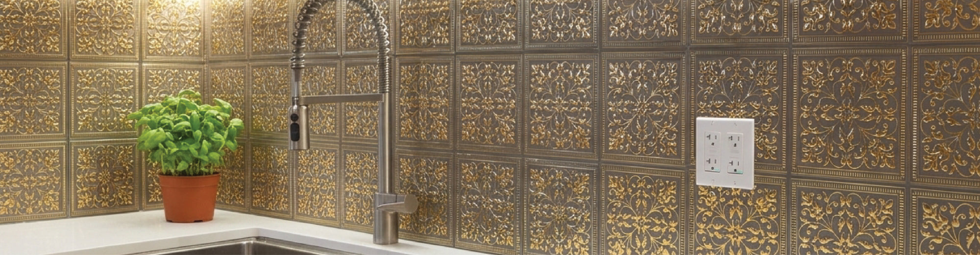Ornate gold and bronze embossed metal tiles covering a kitchen backsplash around a corner, with a tall spring faucet, a basil plant in a terracotta pot and a white double outlet.