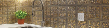 Ornate gold and bronze embossed metal tiles covering a kitchen backsplash around a corner, with a tall spring faucet, a basil plant in a terracotta pot and a white double outlet.
