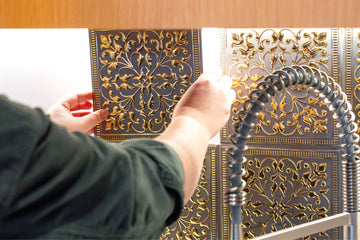 Hands pressing an embossed metal tile into place on a kitchen backsplash, the tiled field ending at the underside of a wood cabinet beside the faucet.