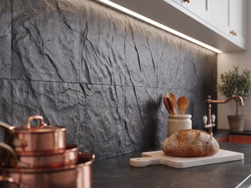 Dark charcoal stone-look backsplash with a rough split-face texture under cabinet lighting, with copper pans, a loaf of bread on a cutting board and wooden spoons on the counter